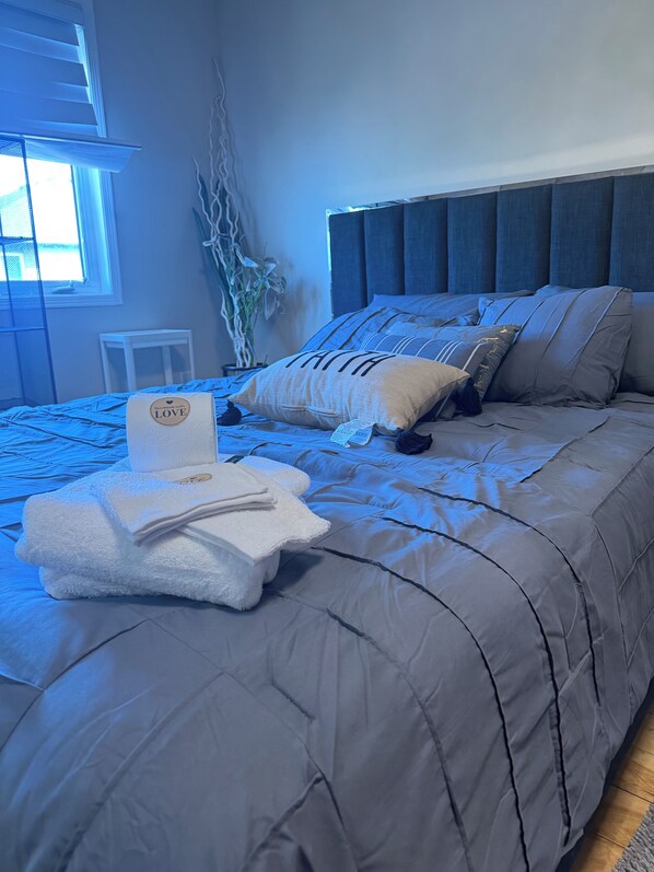 A neatly made bed with a grey duvet, a pillow with the word 'FAITH,' and a folded towel with a logo.