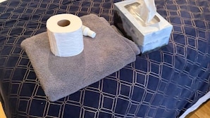 Ein Bett mit einer dunkelblauen, gemusterten Bettdecke, einem weißen Kopfkissen und einer weißen Decke. Auf einem grauen Handtuch auf dem Bett liegen eine Toilettenpapierrolle und eine Papiertuchbox.