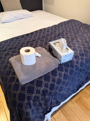 Ein Bett mit einer dunkelblauen, gemusterten Bettdecke, einem weißen Kopfkissen und einer weißen Decke. Auf einem grauen Handtuch auf dem Bett liegen eine Toilettenpapierrolle und eine Papiertuchbox.