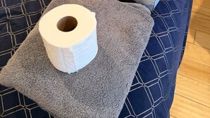 Ein Bett mit einer dunkelblauen Steude, einer Taschentuchbox und einer Toilettenpapierrolle auf einem grauen Kopfkissen.