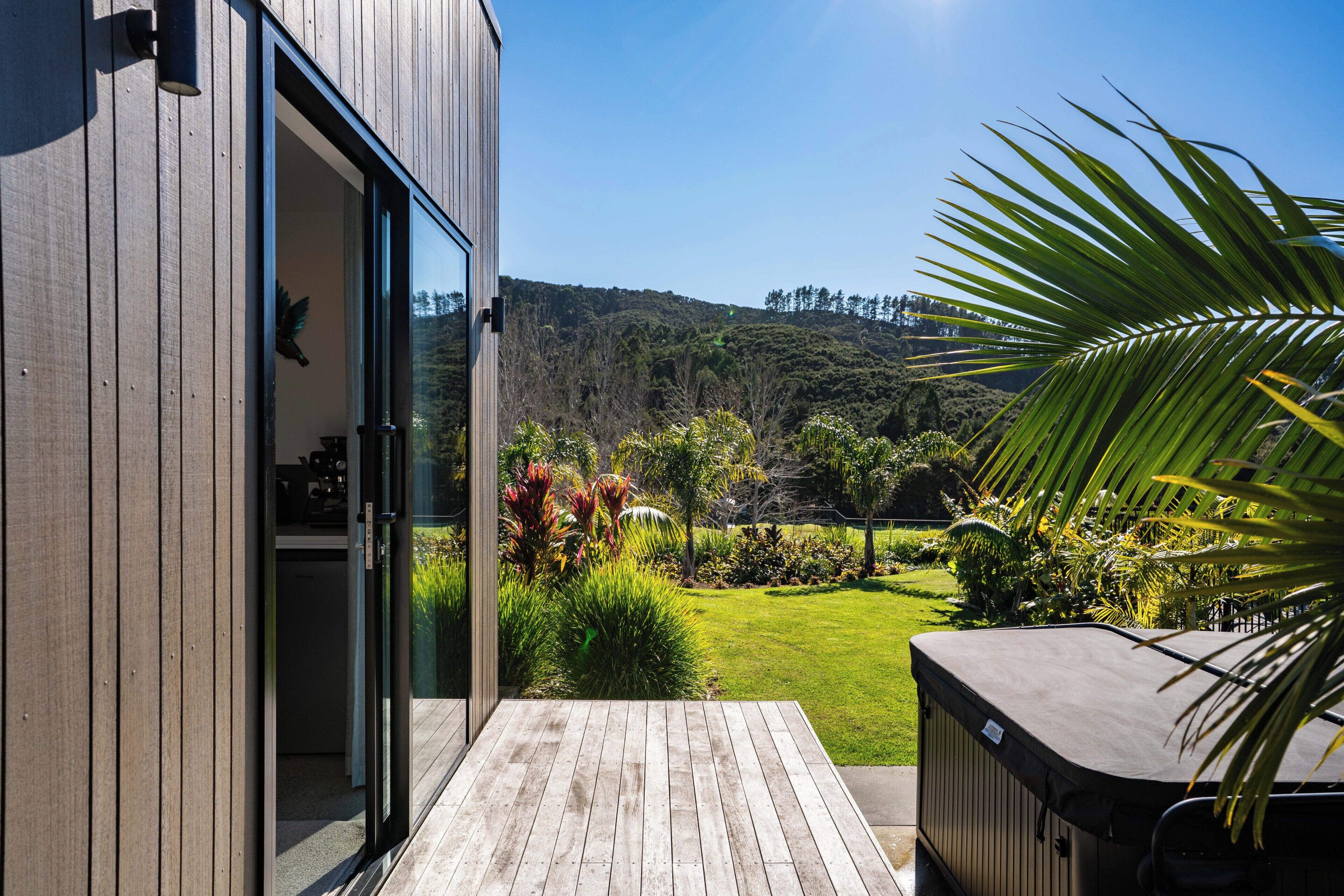 A wooden deck leading to a glass door, with a view of a lush garden and hills in the background.