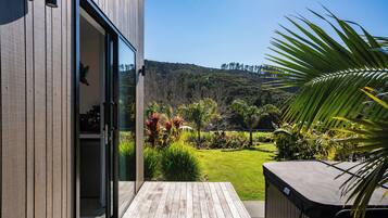 A wooden deck leading to a glass door, with a view of a lush garden and hills in the background.
