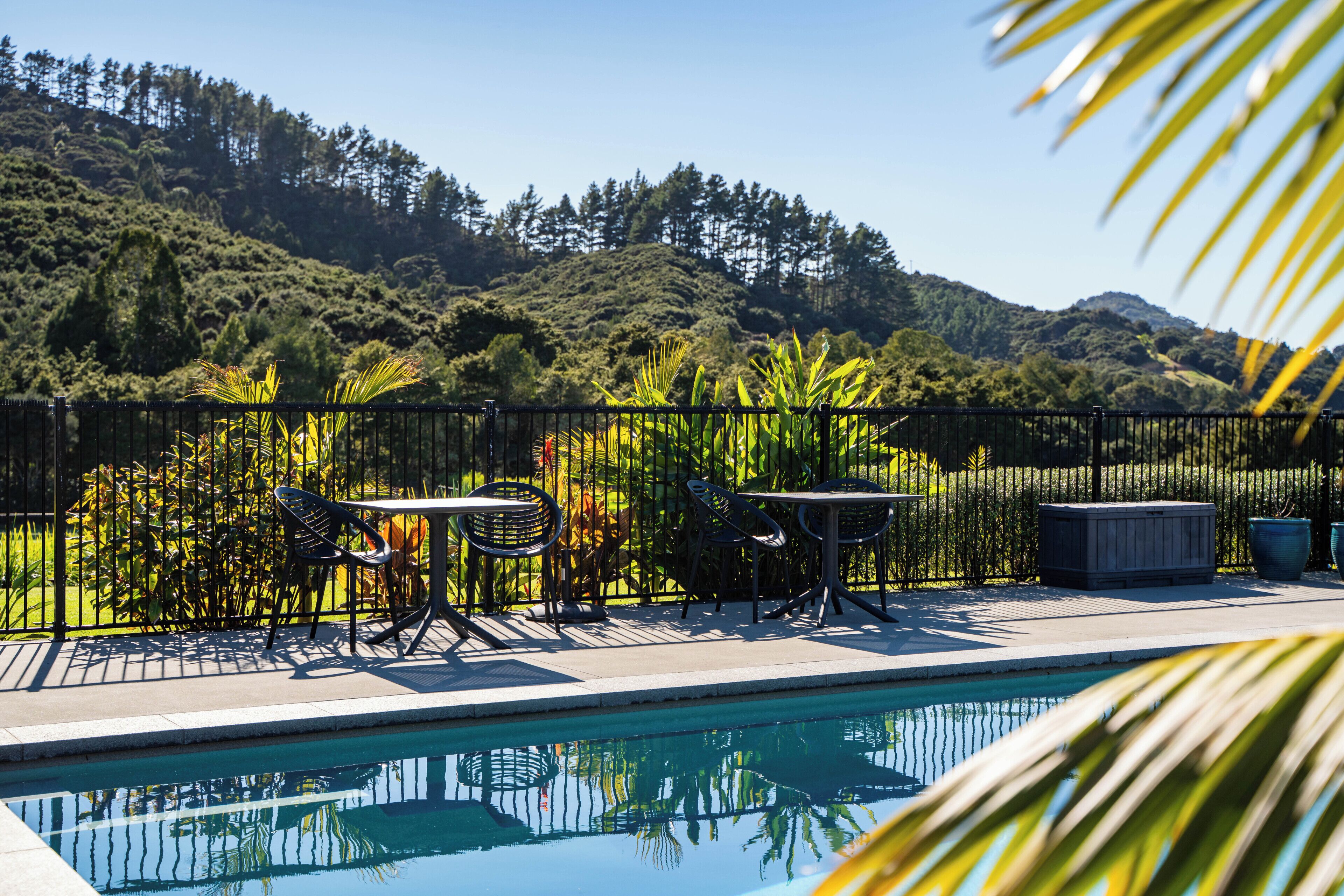 A poolside area with a table and chairs, surrounded by a fence and lush greenery.