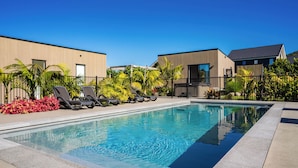 Piscina com espreguiçadeiras e grade, cercada por edifícios e vegetação.