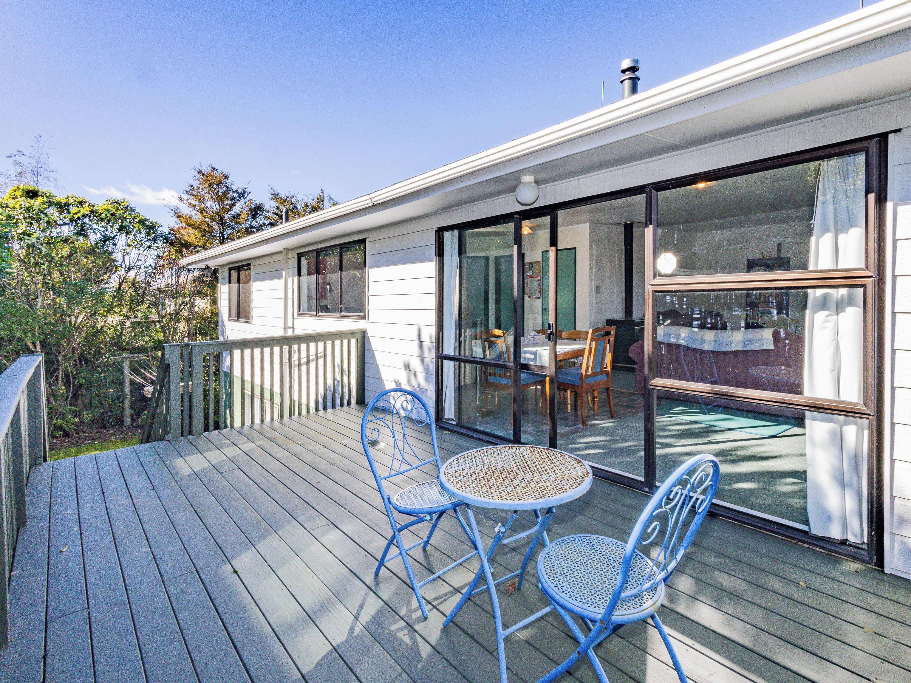 A wooden deck with two blue chairs and a table, a sliding glass door leading to an indoor dining area, and a white building with a chimney.