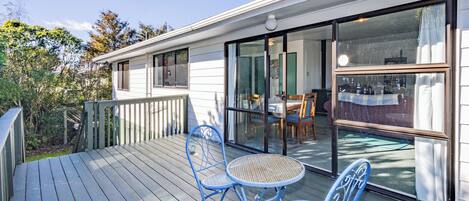 A wooden deck with two blue chairs and a table, a sliding glass door leading to an indoor dining area, and a white building with a chimney.