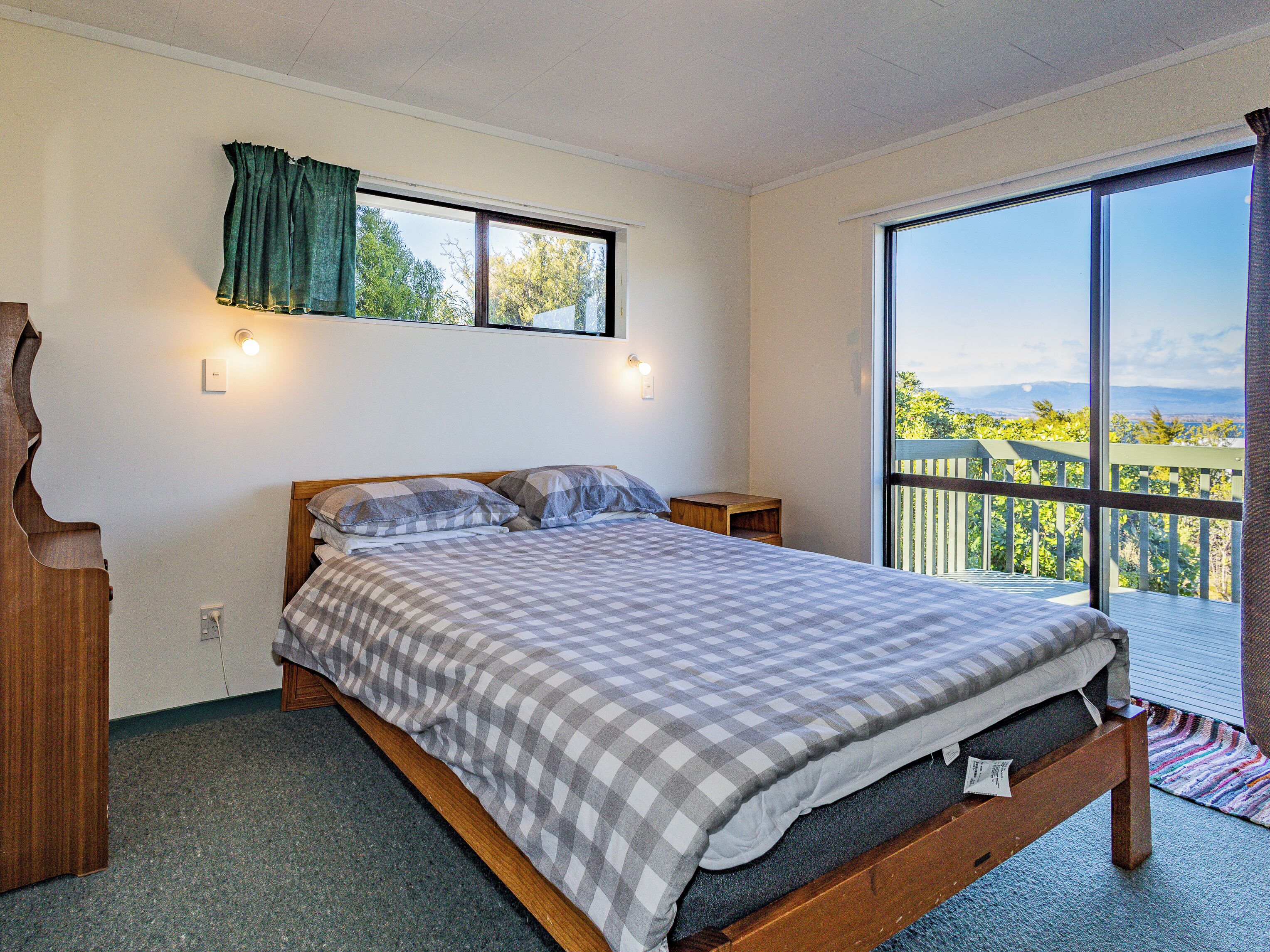 Ein Schlafzimmer mit einem großen Bett, einem Fenster mit Vorhängen, einem Nachttisch und Blick auf einen Balkon mit Geländer.
