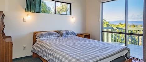 Ein Schlafzimmer mit einem großen Bett, einem Fenster mit Vorhängen, einem Nachttisch und Blick auf einen Balkon mit Geländer.