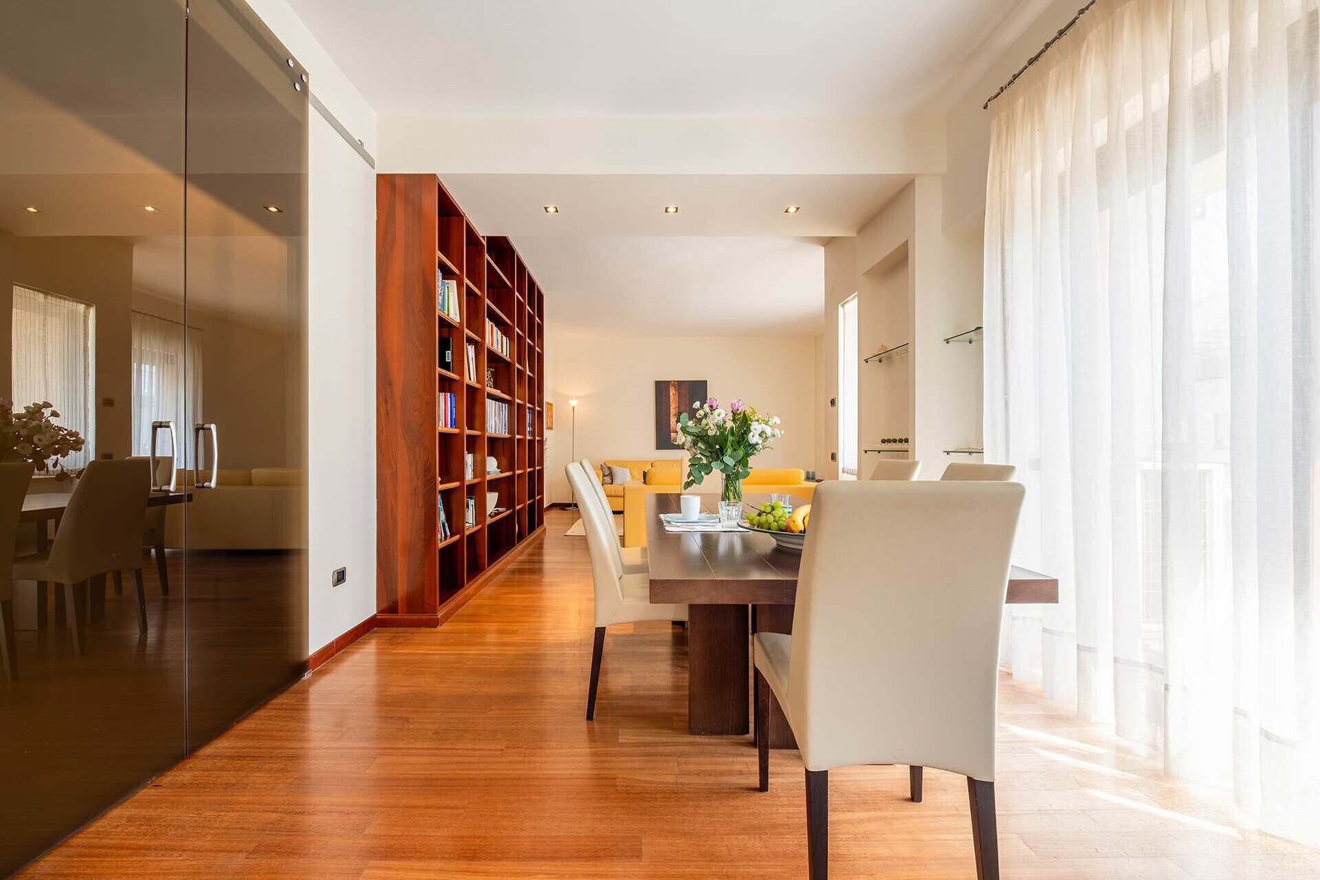 A modern dining area with a wooden bookshelf, a dining table with chairs, and a large window with sheer curtains.