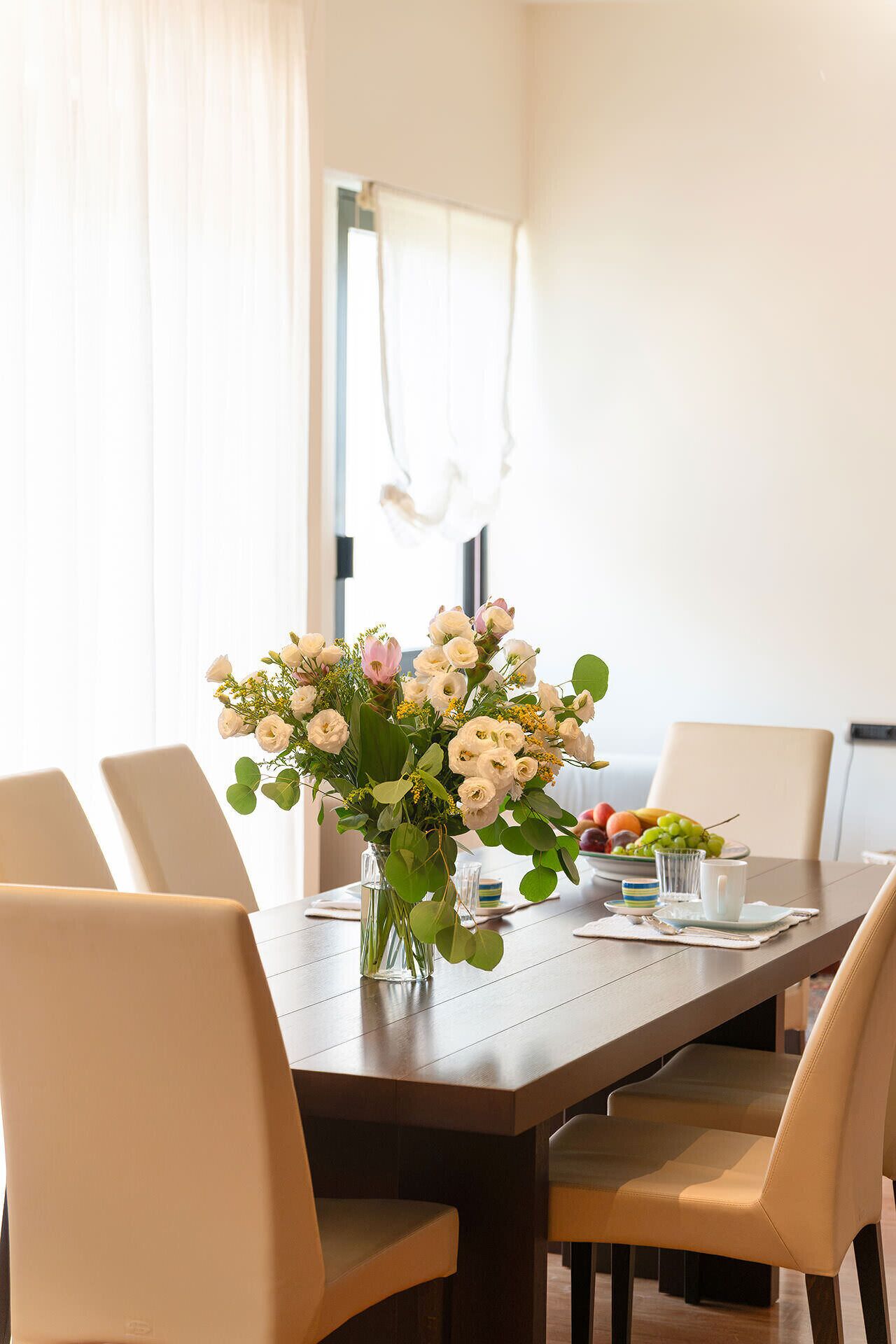 A dining table with chairs, a vase of flowers, and a fruit bowl.