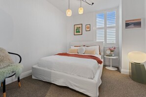 A bedroom with a bed, a chair, a bedside table with a lamp, and a window with blinds. - The Margot by Homello | Designer Apartment | Sky Tower Views | Ponsonby (Freemans Bay)