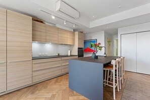 A modern kitchen with wooden cabinets, a central island, and a dining area with chairs. - The Margot by Homello | Designer Apartment | Sky Tower Views | Ponsonby (Freemans Bay)