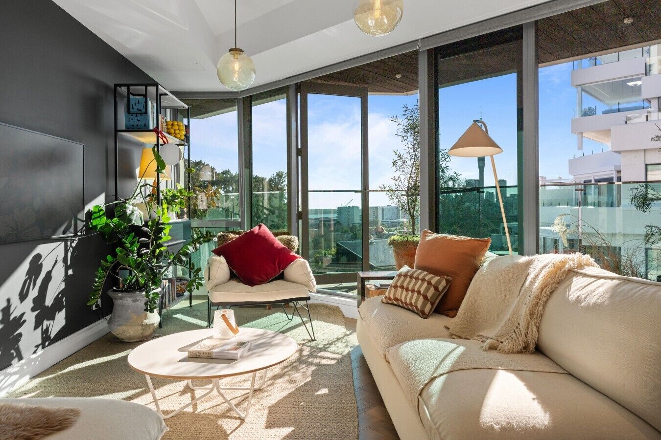 A modern living room with a sofa, armchair, and a round coffee table. There are potted plants and a bookshelf.