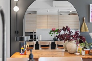 A modern kitchen with wooden cabinets, a dining area with a wooden table, and a large vase with flowers. - The Margot by Homello | Designer Apartment | Sky Tower Views | Ponsonby (Freemans Bay)
