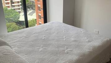Un dormitorio con cama, televisor montado en la pared y ventana con vistas a edificios y árboles.