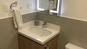 Un baño con un lavamanos blanco, un espejo con iluminación integrada, una toalla colgada en un perchero y un inodoro.