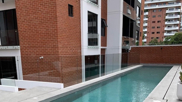 Una piscina rectangular con borde de azulejos, rodeada por un muro de ladrillo y una valla de cristal.