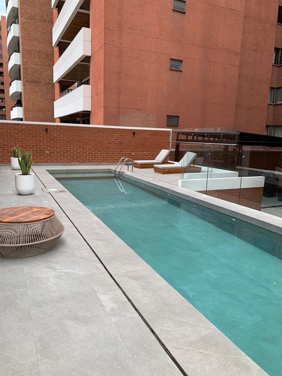 A rooftop pool with lounge chairs and a small table, surrounded by a brick wall and a building in the background.