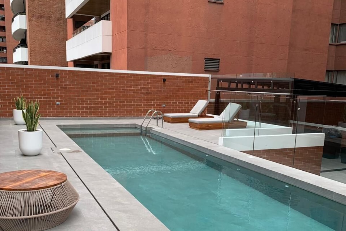 Una piscina en la azotea con sillones de descanso y una pequeña mesa, rodeada por un muro de ladrillo y un edificio al fondo.