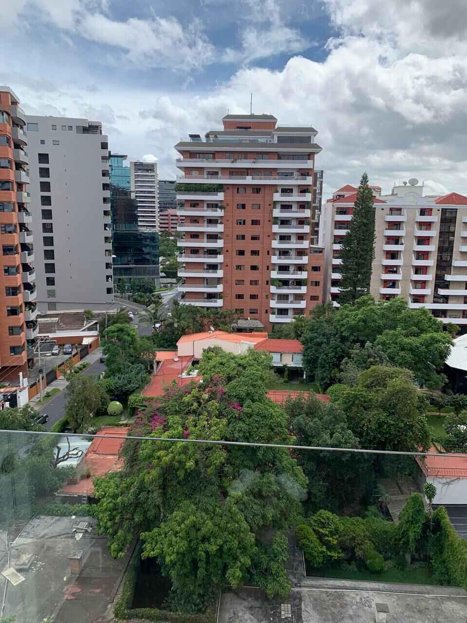 A high-rise residential building with multiple balconies, surrounded by greenery and other buildings.