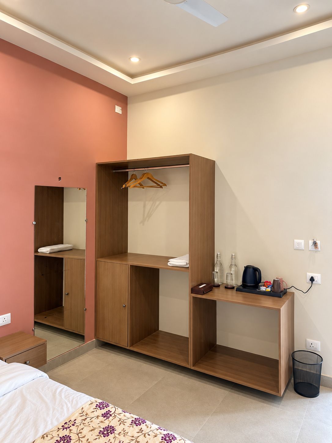 A hotel room with a bed, a wooden wardrobe, a desk with a kettle, and a trash bin.