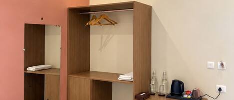 A hotel room with a bed, a wooden wardrobe, a desk with a kettle, and a trash bin.