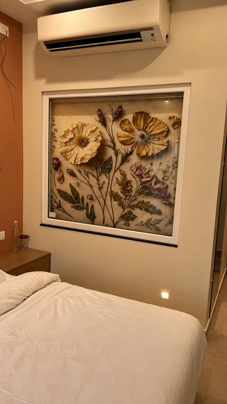 A bedroom with a bed, an air conditioning unit, and a framed floral artwork on the wall.