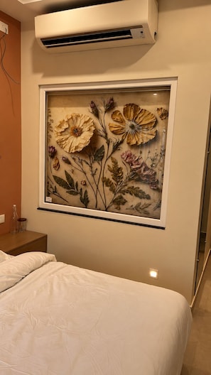 A bedroom with a bed, an air conditioning unit, and a framed floral artwork on the wall.