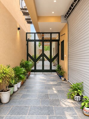 A narrow outdoor corridor with a tiled floor, flanked by potted plants and a closed roller shutter on one side.