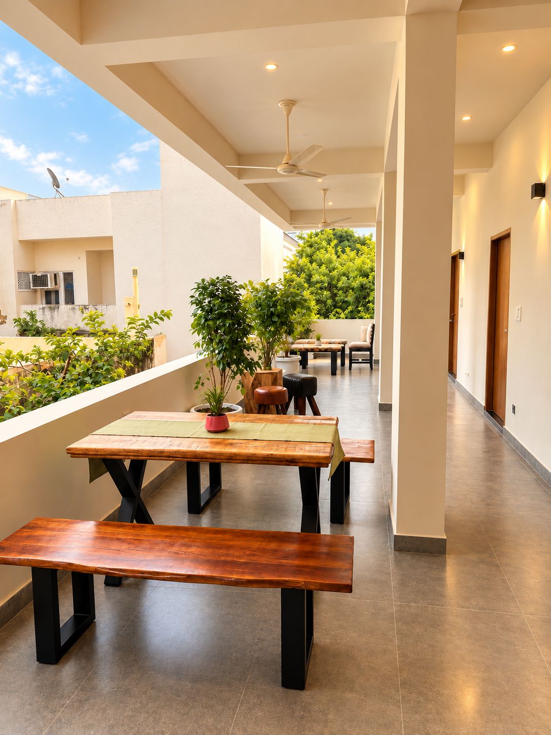 Un vaste couloir avec un banc et une table en bois, un ventilateur de plafond et une vue sur la verdure à l’extérieur.