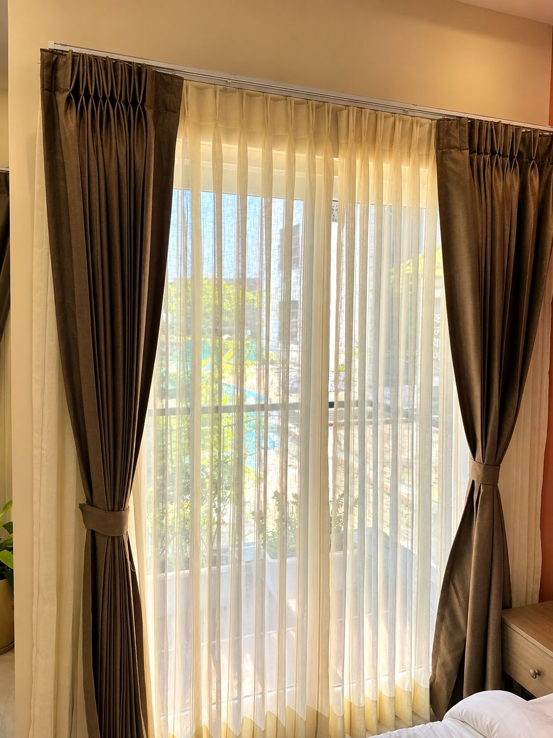 Una habitación con un ventanal grande, cortinas traslúcidas recogidas, que ofrece vistas a zonas verdes y un edificio.