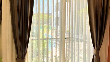 Una habitación con un ventanal grande, cortinas traslúcidas recogidas, que ofrece vistas a zonas verdes y un edificio.