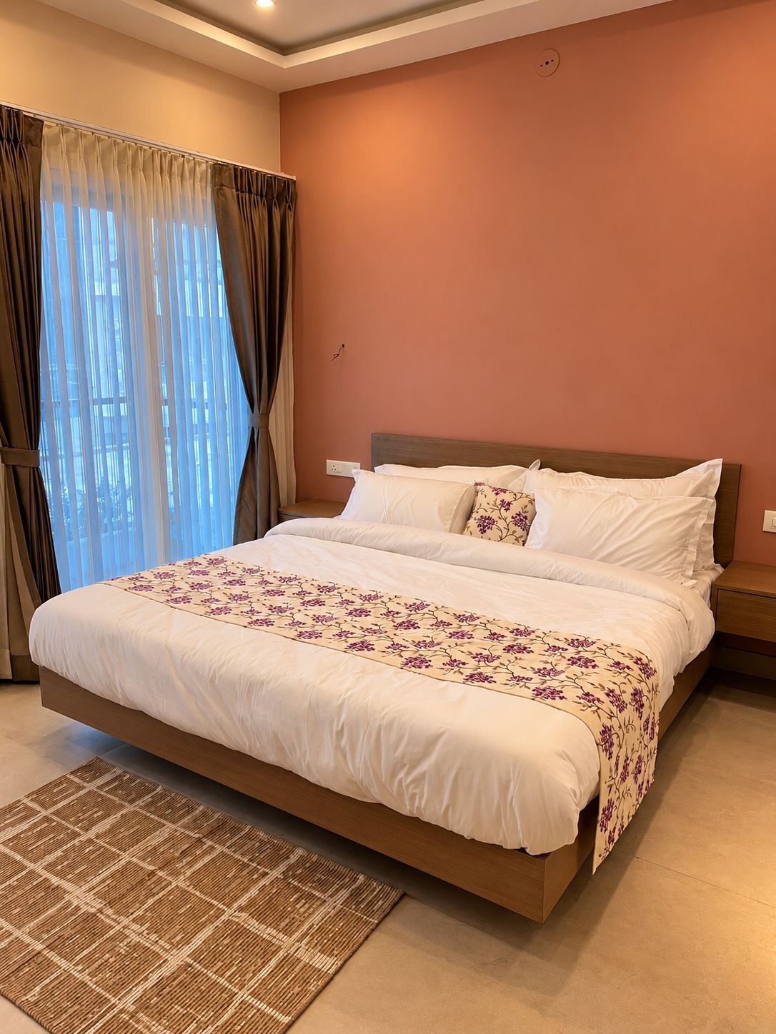 Una habitación de hotel con una cama grande, una mesita de noche, cortinas en la ventana y una lámpara de pared.