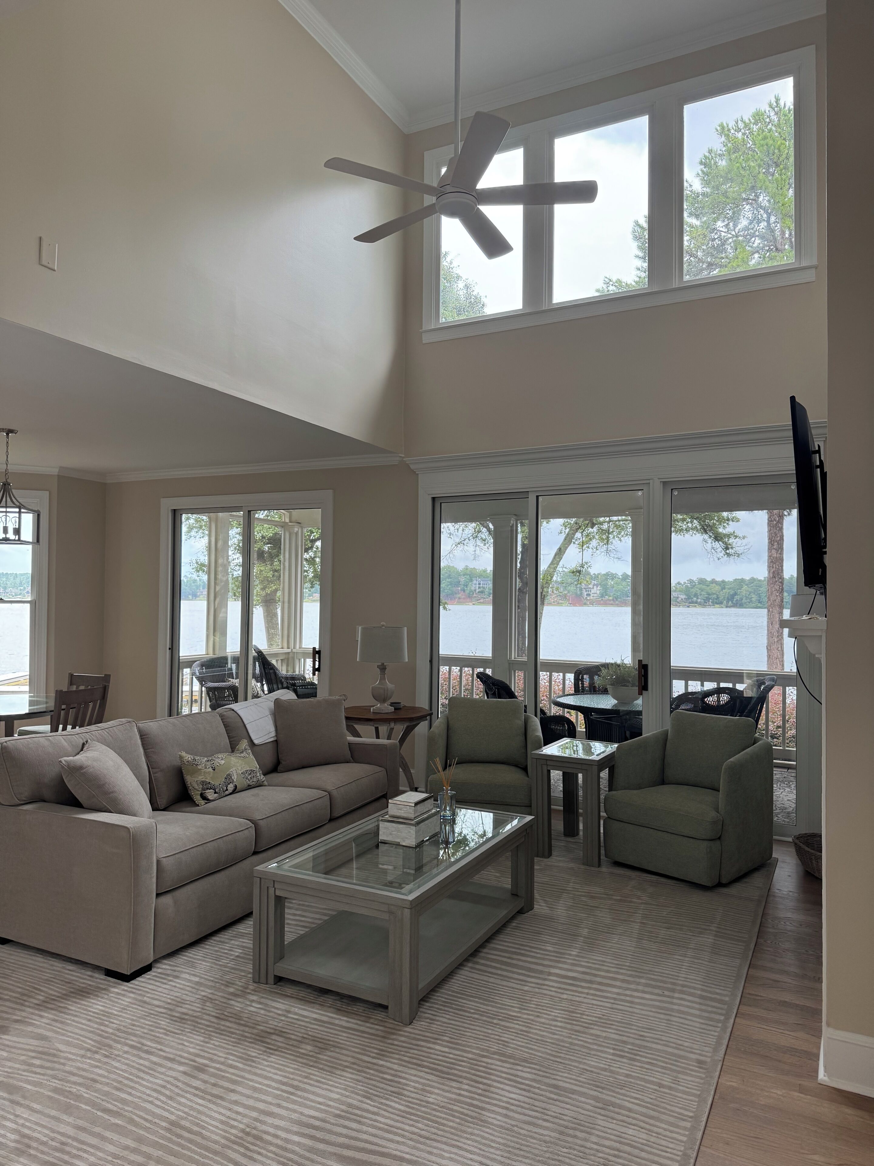 A spacious living room with a high ceiling, large windows, a ceiling fan, and a view of a body of water.