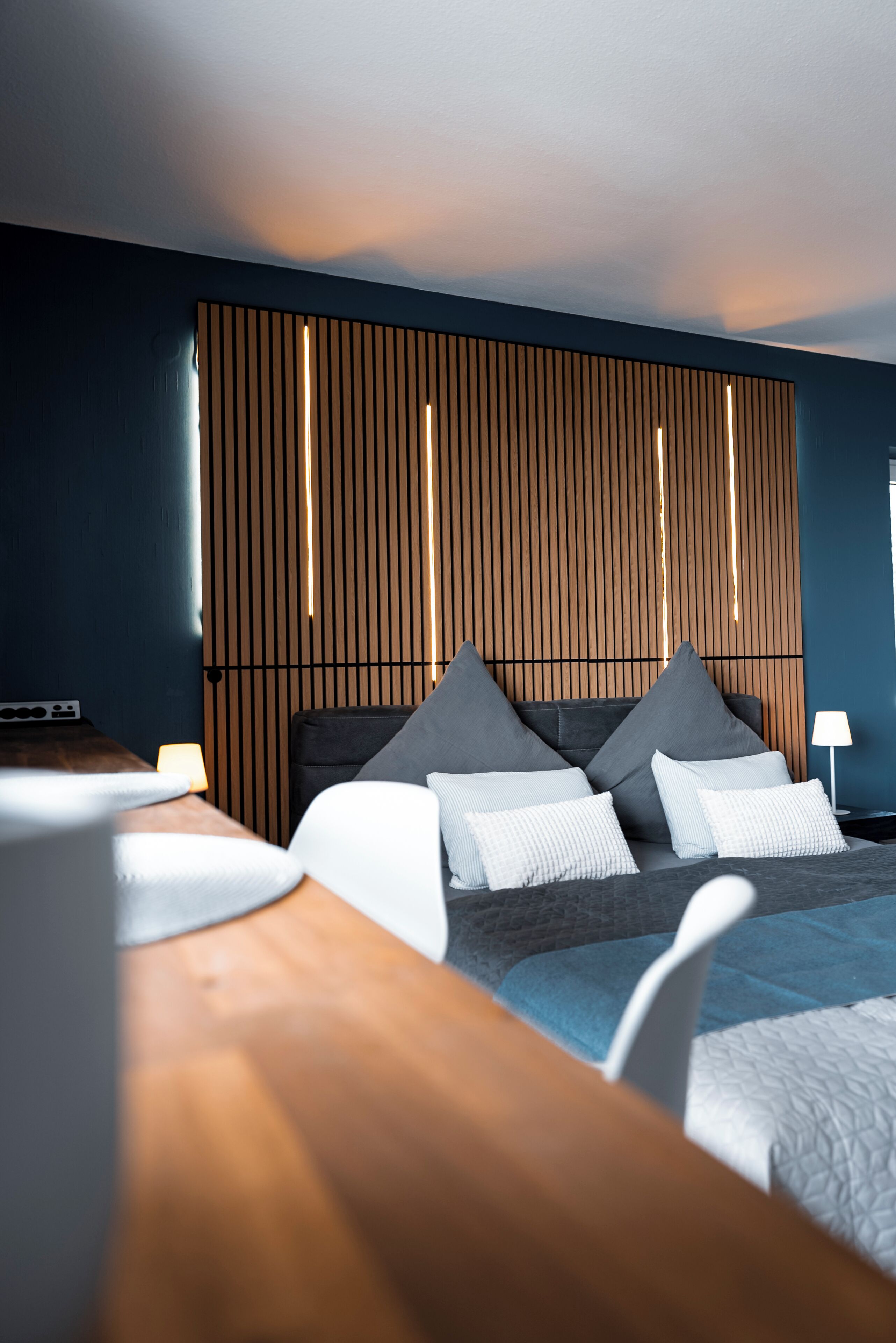 Ein modernes Hotelzimmer mit einem hölzernen Kopfteil, einem Bett mit grau-weißer Bettwäsche, einem Holztisch und einem Nachttisch mit einer Lampe.