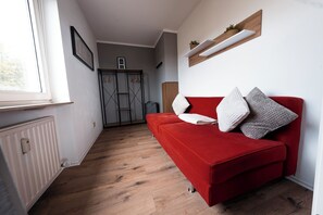A red sofa with pillows, a radiator, a wooden floor, a window, and a door with a coat rack. - Vrbo Property (Linz am Rhein)
