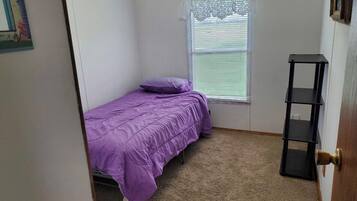 Quarto pequeno com uma cama de solteiro, janela com persianas, porta e uma estante preta.