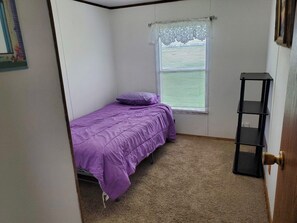 A small bedroom with a single bed, a window with blinds, a door, and a black shelf.
