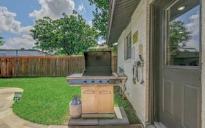 An outdoor grill with four burners, a gas tank, and a wooden cabinet, located in a backyard with a wooden fence and a house in the background. - Modern NRH Retreat | Pool, BBQ & Sleeps 10 (Fort Worth)