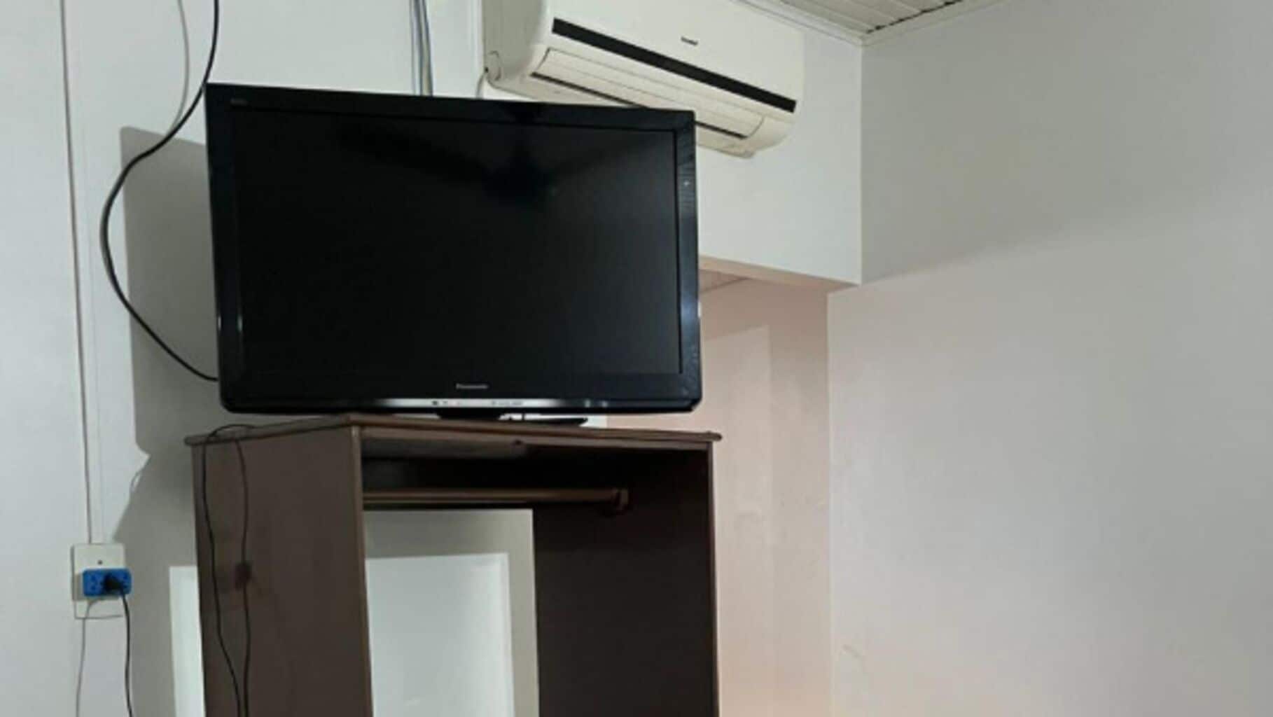 Televisão sobre suporte em um quarto com paredes brancas e um ar condicionado acima.