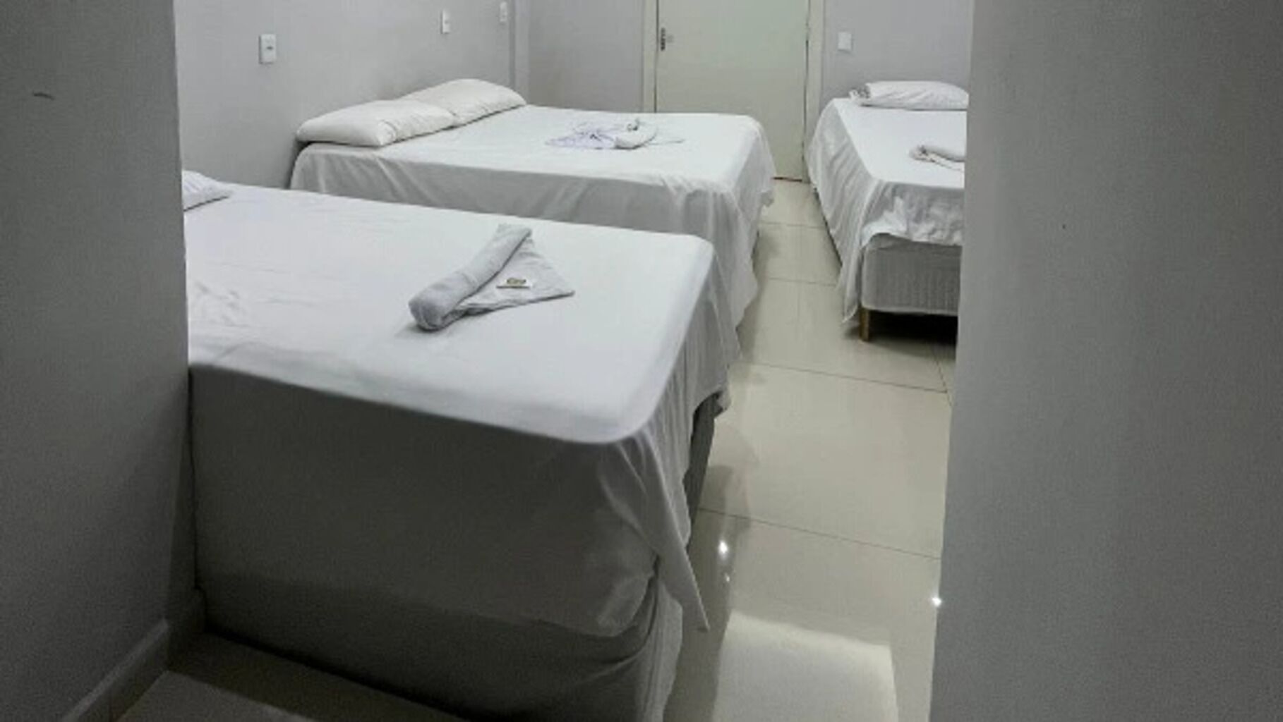 A corridor with three single beds, each with a white bedspread and a pillow. The room has a tiled floor and white walls.