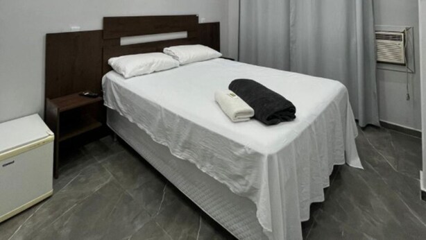 Uma cama de solteiro com um cobreleito branco, duas almofadas, uma toalha preta e uma mesa de cabeceira com abajur.