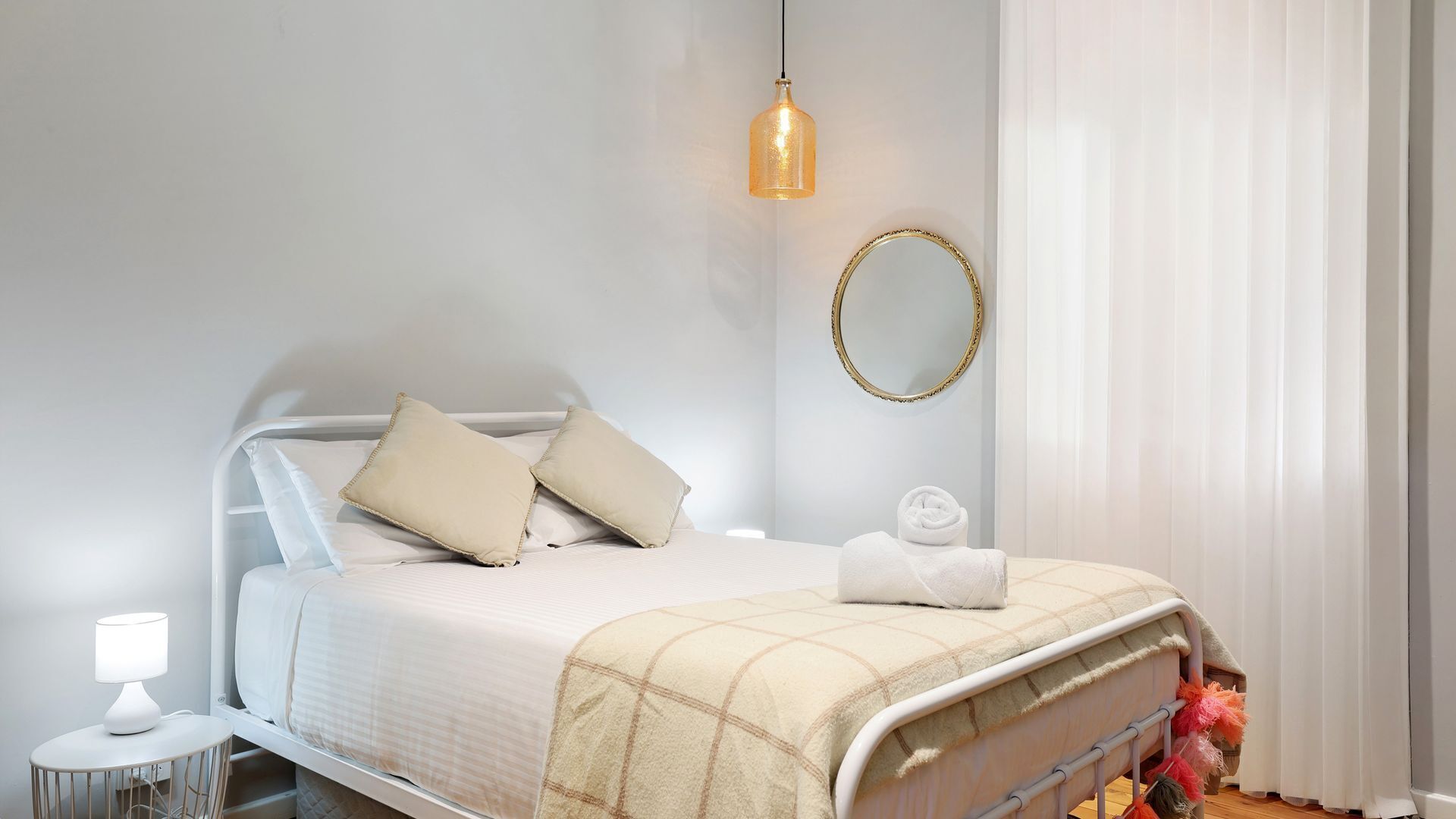A neatly made bed with a white and beige checkered blanket, a bedside lamp, and a round mirror on the wall.