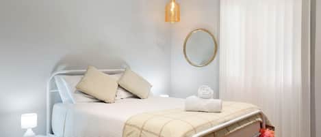 A neatly made bed with a white and beige checkered blanket, a bedside lamp, and a round mirror on the wall.