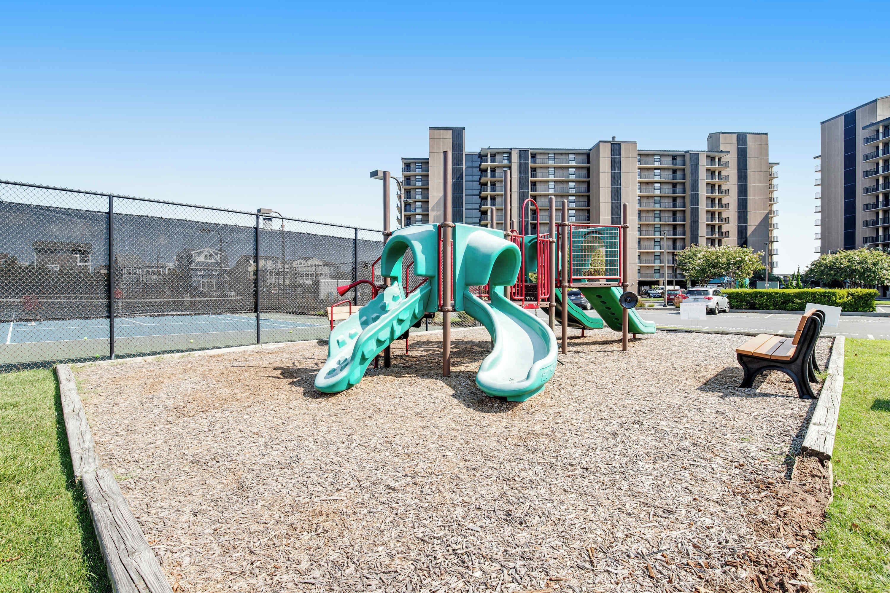 A playground with slides and climbing structures, surrounded by a fence and residential buildings in the background.