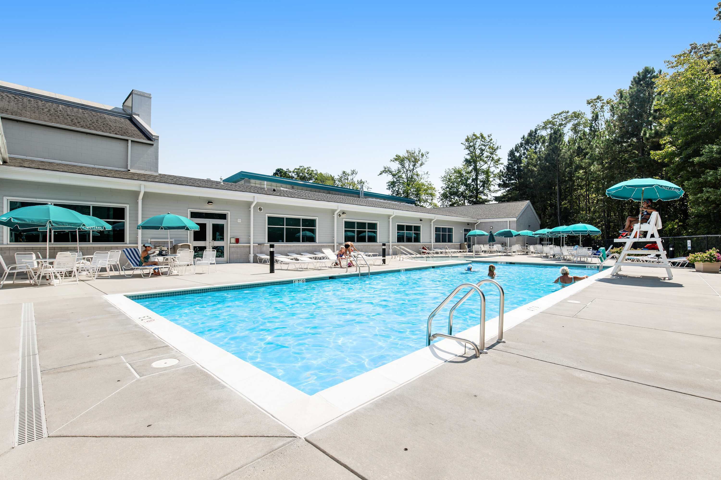 Une piscine avec des chaises longues et des parasols, un poste de surveillance pour les sauveteurs, et un bâtiment doté de grandes fenêtres.