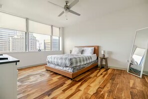 Een slaapkamer met een bed, een plafondventilator, een raam met uitzicht op gebouwen en een houten vloer.