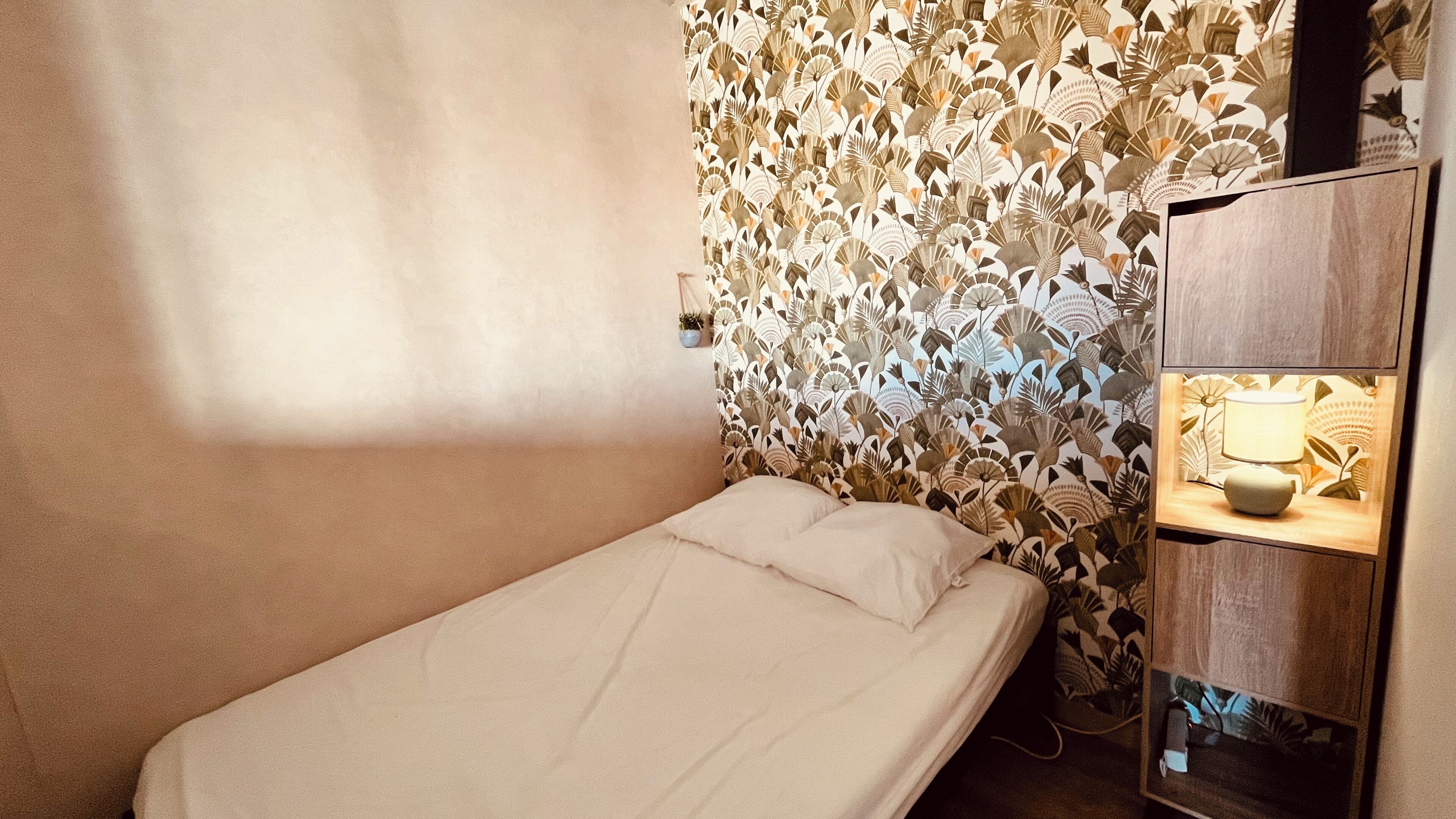 A single bed with a plain mattress and pillows, a wooden nightstand with a lamp, and a wall with a leaf-patterned wallpaper.