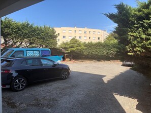 Ein Parkplatz mit einem schwarzen Auto und einem blauen Fahrzeug, umgeben von Bäumen und einem Gebäude im Hintergrund.
