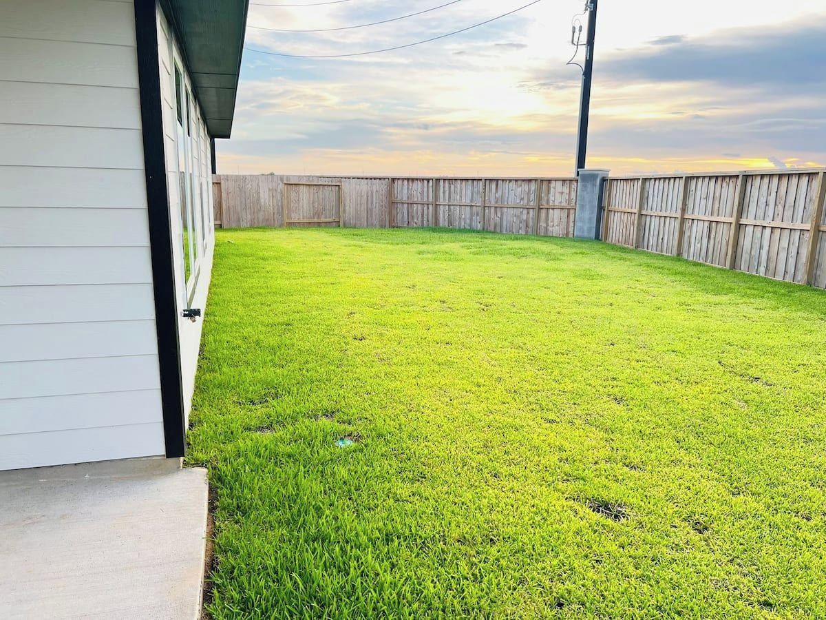 A backyard with a wooden fence, a grassy lawn, and a house with a white exterior.